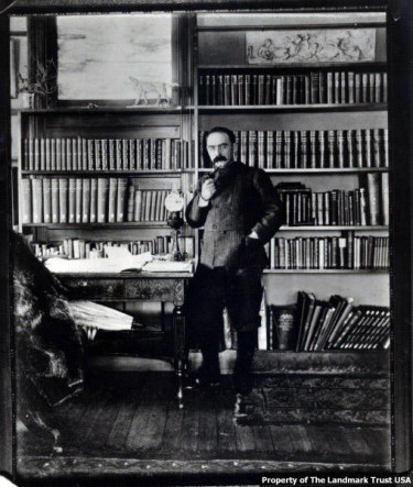 Rudyard Kipling holds and pipe in his hand and stands next to a table in fornt of shelves filled with books