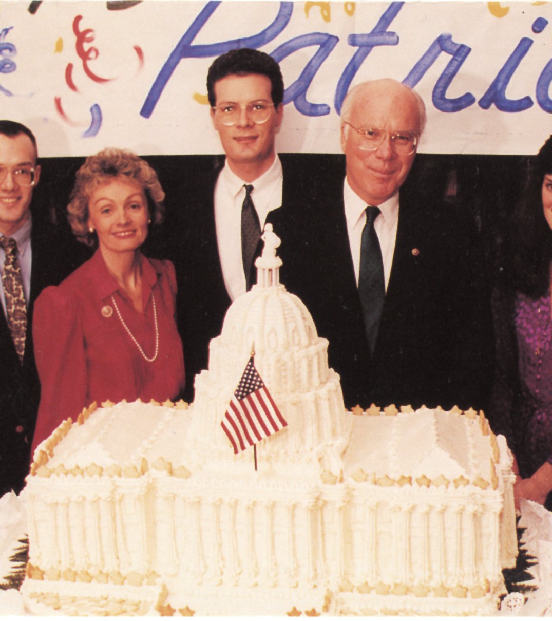 Marcelle Leahy in a purple dress, her son in a suit, and Senator Patrick Leahy in a suit stand behind a cake shaped like the U.S. Capitol building.
