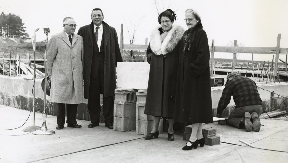 Two men and two women in winter coats stand in front of a building construction site next to a microphone, and to their right, a workman in a plain jacket kneels on the concrete.